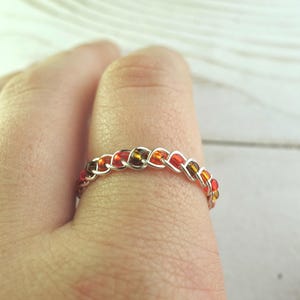 May include: A silver-toned ring with a chain-like design. The ring is adorned with small, colorful beads in shades of red, orange, and amber. The ring is worn on a finger, showcasing the intricate beadwork and metallic sheen.