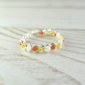 Può includere: Un anello in filo d'argento con un design intrecciato. L'anello è adornato con piccole perline colorate bianche, gialle e arancioni. L'anello è posto su una superficie di legno chiaro.
