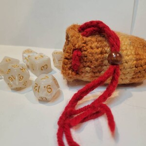 May include: A crocheted dice bag with a red drawstring and a wooden toggle. The bag is filled with white dice with gold numbers.