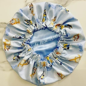 Puede incluir: Un gorro forrado de sat&eacute;n azul claro con un patr&oacute;n repetido de perros de dibujos animados. El gorro tiene un borde fruncido y el texto "Melody Rose Boutique" est&aacute; bordado en el forro de sat&eacute;n.