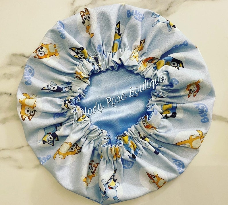 Puede incluir: Un gorro forrado de sat&eacute;n azul claro con un patr&oacute;n repetido de perros de dibujos animados. El gorro tiene un borde fruncido y el texto "Melody Rose Boutique" est&aacute; bordado en el forro de sat&eacute;n.