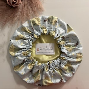 May include: A light blue satin shower cap with a print of a cartoon princess in a yellow dress. The cap has a yellow satin lining and the text "Forever Stitched" and "www.foreverstitched.com" printed on the lining.