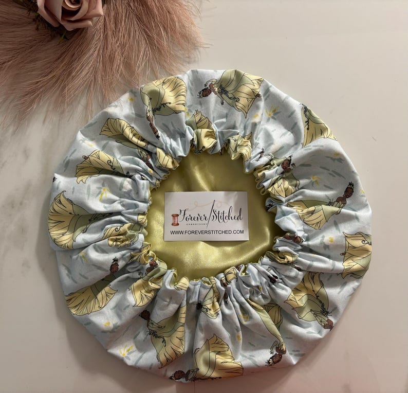 May include: A light blue satin shower cap with a print of a cartoon princess in a yellow dress. The cap has a yellow satin lining and the text "Forever Stitched" and "www.foreverstitched.com" printed on the lining.