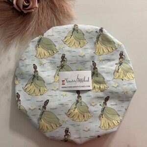 May include: A light blue satin shower cap with a repeating pattern of a cartoon princess in a green dress and a crown. The princess is standing on a lily pad with white flowers. The text "Forever Stitched" and "www.foreverstitched.com" is printed on a white tag.