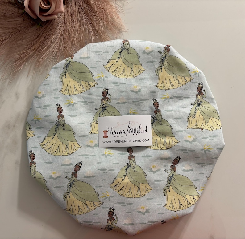 May include: A light blue satin shower cap with a repeating pattern of a cartoon princess in a green dress and a crown. The princess is standing on a lily pad with white flowers. The text "Forever Stitched" and "www.foreverstitched.com" is printed on a white tag.