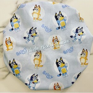 Puede incluir: Un art&iacute;culo de tela azul claro con un patr&oacute;n repetido de perros de dibujos animados en c&iacute;rculos blancos. La palabra "Bluey" est&aacute; impresa en azul claro. El texto "Melody Rose Boutique" tambi&eacute;n es visible.