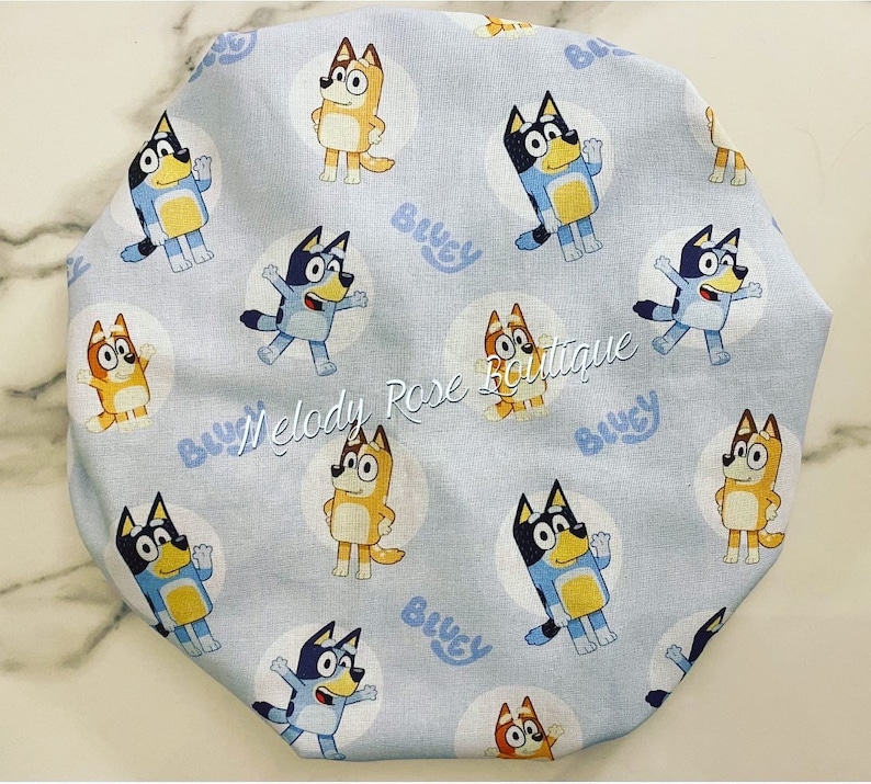 Puede incluir: Un art&iacute;culo de tela azul claro con un patr&oacute;n repetido de perros de dibujos animados en c&iacute;rculos blancos. La palabra "Bluey" est&aacute; impresa en azul claro. El texto "Melody Rose Boutique" tambi&eacute;n es visible.
