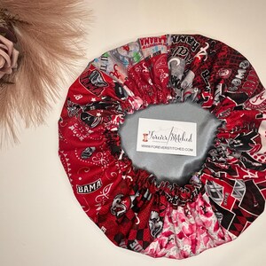 May include: Red, white, and black satin bonnet with a patchwork design featuring various college logos and the words "BAMA" and "ROLL TIDE". The bonnet is lined with a gray satin fabric and has a tag that reads "Forever Stitched".