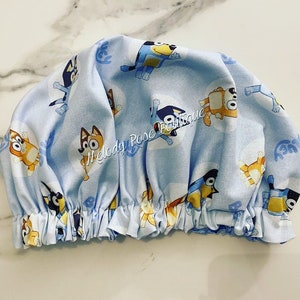 Puede incluir: Un gorro de ducha azul claro con un estampado repetido de perros de dibujos animados en varias poses. El gorro tiene un borde el&aacute;stico fruncido. El texto "Melody Rose Boutique" es visible en el gorro.