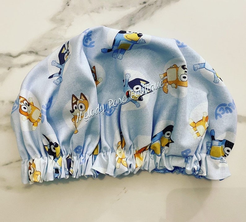 Puede incluir: Un gorro de ducha azul claro con un estampado repetido de perros de dibujos animados en varias poses. El gorro tiene un borde el&aacute;stico fruncido. El texto "Melody Rose Boutique" es visible en el gorro.