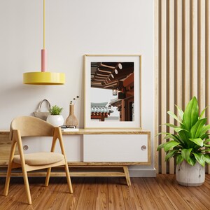 May include: A framed print of a traditional Asian building with a wooden roof and a lantern hanging from the eaves. The building is painted in shades of brown and red, and the roof is covered in blue tiles. The print is hanging on a white wall above a wooden cabinet.