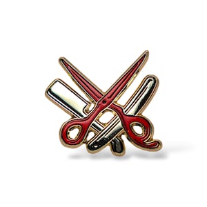 May include: A gold-colored barber pin featuring a red pair of scissors, a straight razor, and a barber pole design. The scissors have red blades and a gold outline. The straight razor is silver with a gold outline.
