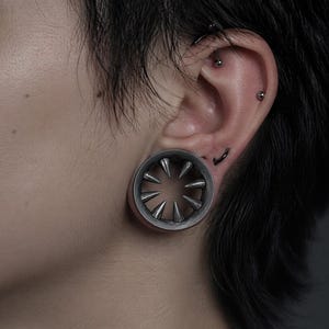 Puede incluir: Un dilatador de oreja circular de metal oscuro con una serie de púas orientadas hacia el interior, dispuestas en un patrón radial alrededor de la abertura central, insertado en un lóbulo estirado con otros pequeños piercings plateados.