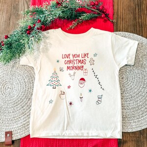 May include: Off-white short-sleeved t-shirt with the text "Love You Like Christmas Morning" in red. The shirt features Christmas-themed illustrations in blue, red, and green, including a Christmas tree, Santa, and a stocking.