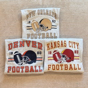 Op de afbeelding: Drie grijze sweatshirts met voetbalontwerpen. Elk shirt heeft een voetbal, een helm en het woord "FOOTBALL" samen met een stadsnaam: New Orleans, Denver en Kansas City. De shirts tonen ook de nummers 18, 87, 80 en 93.