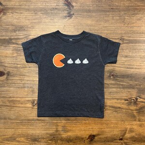 May include: A dark gray t-shirt with a Pac-Man design featuring a slice of pumpkin pie eating three white marshmallows.
