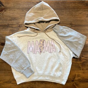 May include: A light gray hooded sweatshirt with a brown hood. The sleeves are gray and the front of the sweatshirt has the word "MAMA" printed in pink with a floral lightning bolt design.