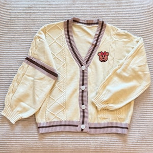 May include: Cream-colored cardigan with a V-neck, featuring a cable knit design on the sleeves and front panels. The sweater has brown and light purple stripes along the edges and a cartoon character patch on the chest. It has four white buttons.