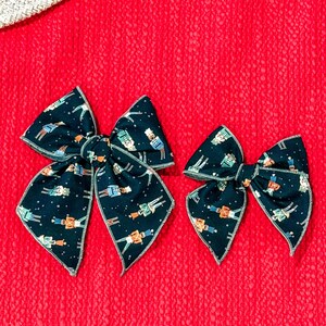 May include: Two dark blue fabric hair bows with a repeating pattern of toy soldiers and stars. The bows are on a textured red background. One bow is larger than the other.
