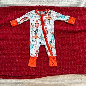 May include: A white baby sleeper with a festive print featuring ballerinas, nutcrackers, and candy canes. The long-sleeved onesie has orange cuffs and a red zipper down the front. The sleeper is laying on a red textured surface.