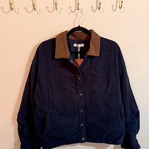 May include: A navy blue corduroy jacket featuring a brown ribbed collar, long sleeves, and a front button closure with metallic buttons. The garment includes two chest pockets and is displayed on a black hanger against a plain wall.