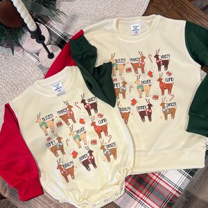 May include: Two cream-colored long-sleeved shirts with red and green sleeves. The shirts feature a festive design of reindeer with the names of Santa's reindeer printed on them. The shirts are a fun and festive way to celebrate the holiday season.