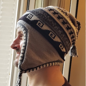 Może przedstawiać: Brązowa i biała dzianinowa czapka z długim, splecionym pomponem. Czapka ma geometryczny wzór i klapkę, którą można opuścić, aby zakryć uszy.