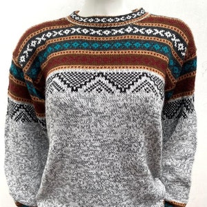 Peut inclure: Un pull en tricot gris avec un motif géométrique coloré autour du cou et des poignets. Le motif est composé de couleurs brun, rouge, bleu et turquoise.