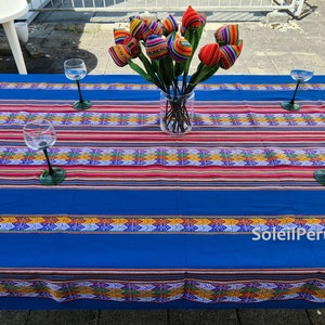 Puede incluir: Un mantel azul con rayas de colores y patrones geométricos. Cuatro copas de vino están colocadas sobre la mesa, y un jarrón con un ramo de flores de tela de colores está en el centro. El texto "Soleil Peruvien" está impreso en el mantel.