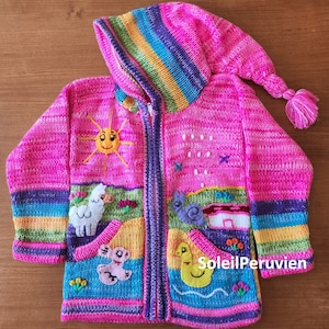 May include: A pink knitted hooded sweater with colorful stripes and embroidered details, including a sun, a llama, a bird, a house, and a duck. The sweater has a zipper closure and two pockets. The text "Soleil Peruvien" is embroidered on the bottom right pocket.