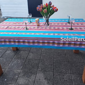 Peut inclure: Une nappe rayée bleue et rouge avec une bordure à motif géométrique coloré. La nappe est posée sur une table en bois avec quatre verres à vin et un vase de tulipes colorées. Le texte "SoleilPeruvien" est visible dans le coin inférieur droit de l'image.
