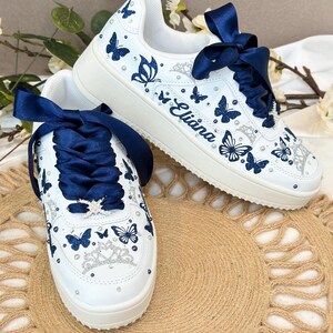 Puede incluir: Zapatillas blancas adornadas con dise&ntilde;os de mariposas azul marino y detalles brillantes. Los zapatos tienen cordones de cinta de raso azul marino y el nombre "Eliana". Una corona decorativa est&aacute; en la puntera.