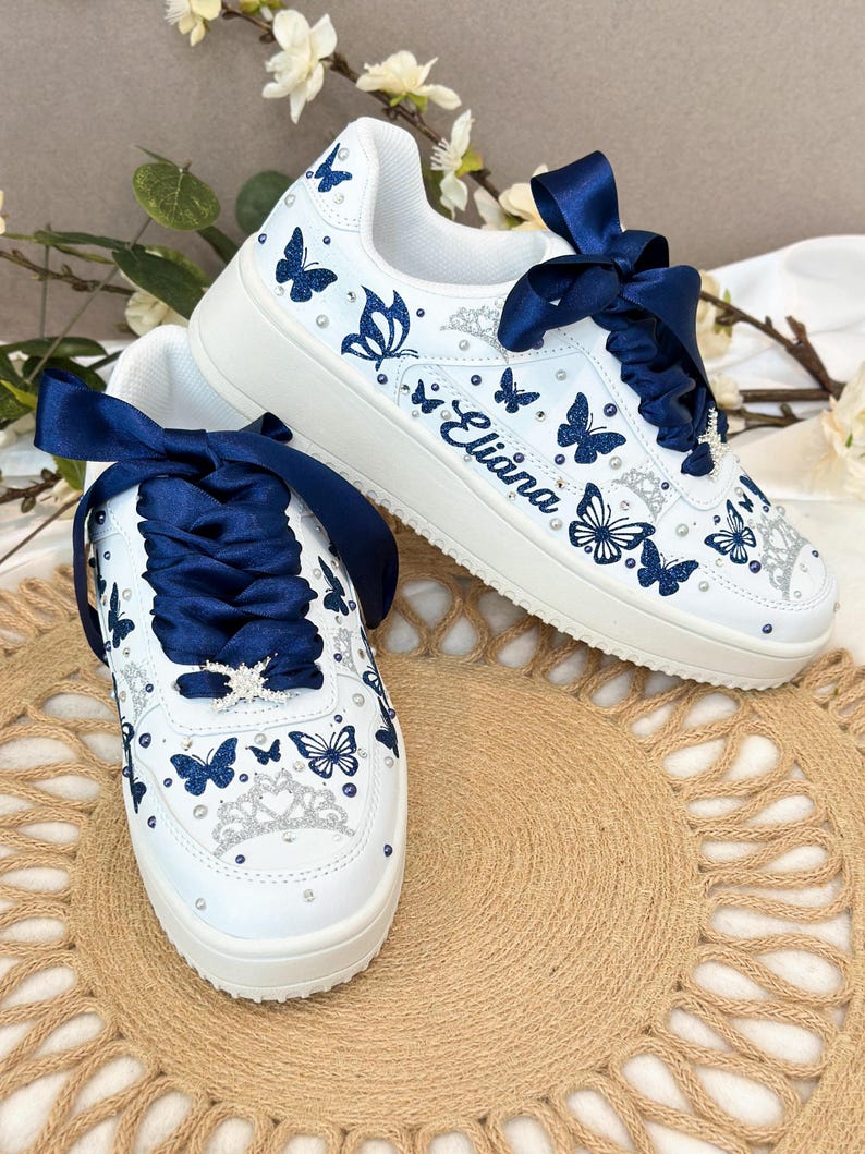 Puede incluir: Zapatillas blancas adornadas con dise&ntilde;os de mariposas azul marino y detalles brillantes. Los zapatos tienen cordones de cinta de raso azul marino y el nombre "Eliana". Una corona decorativa est&aacute; en la puntera.