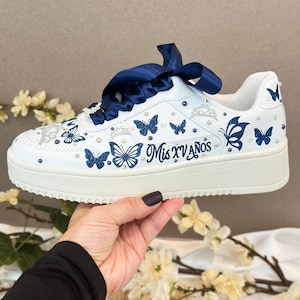 Puede incluir: Zapatillas blancas con dise&ntilde;os de mariposas azules y el texto "Mis XV A&ntilde;os". Los zapatos tienen cordones de cinta de sat&eacute;n azul y est&aacute;n decorados con peque&ntilde;as perlas. Una opci&oacute;n de calzado &uacute;nica y elegante.