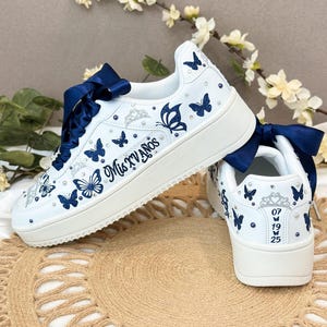 Puede incluir: Zapatillas blancas decoradas con mariposas azul marino y pedrer&iacute;a. Los zapatos presentan lazos azul marino, el texto "Mis XV A&ntilde;os" y la fecha "07 19 25". Estas zapatillas personalizadas son perfectas para una ocasi&oacute;n especial.