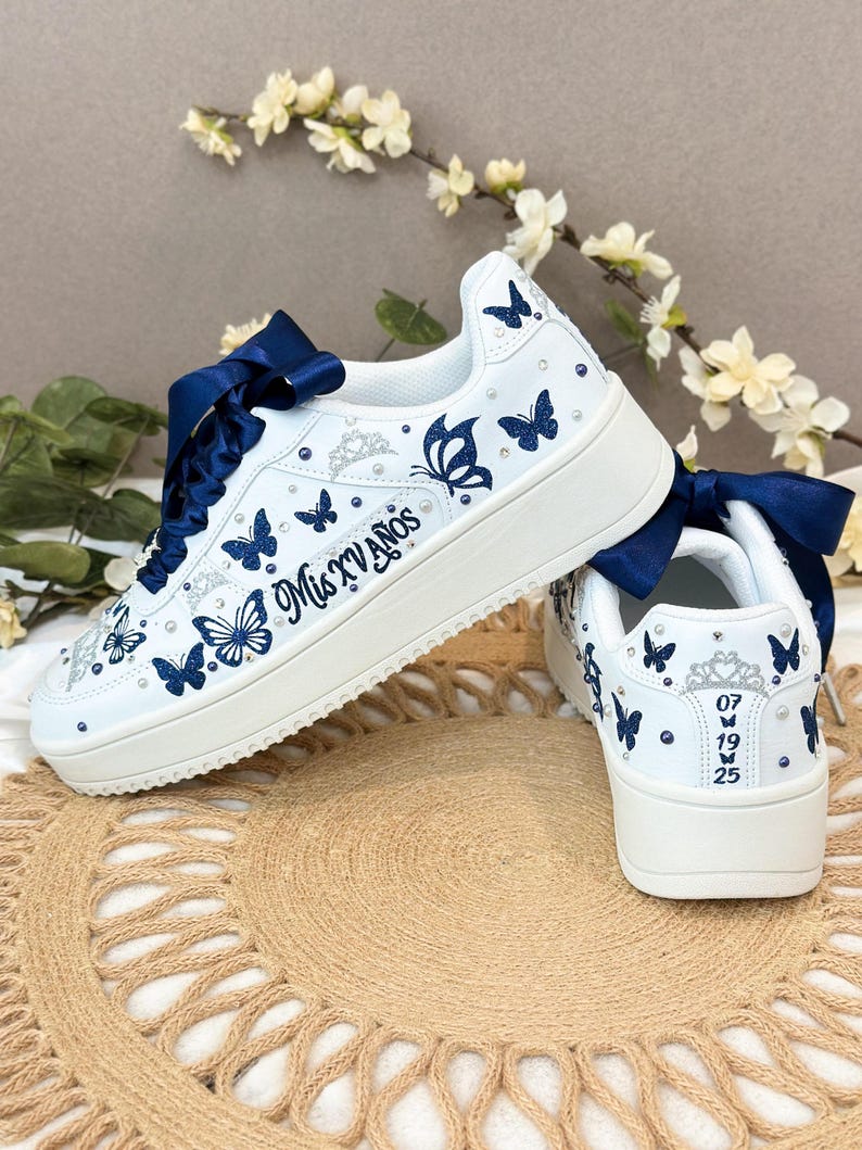 Puede incluir: Zapatillas blancas decoradas con mariposas azul marino y pedrer&iacute;a. Los zapatos presentan lazos azul marino, el texto "Mis XV A&ntilde;os" y la fecha "07 19 25". Estas zapatillas personalizadas son perfectas para una ocasi&oacute;n especial.