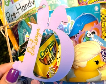 May include: A pink wooden letter B with the name "Da'Laya" written in gold script. The letter is in a basket with other Easter basket items, including a box of Crayola crayons, a book, and a yellow rubber ducky.
