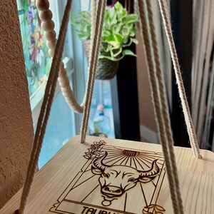 May include: A wooden wall hanging with a rope macrame design. The hanging features a wood-burned image of the Taurus zodiac sign with the word "Taurus" below it.