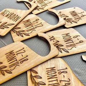 May include: Wooden hangers with engraved text indicating different age ranges for babies, such as "Newborn", "0-3 Months", "3-6 Months", "6-9 Months", "9-12 Months", "12-18 Months", and "18-24 Months".