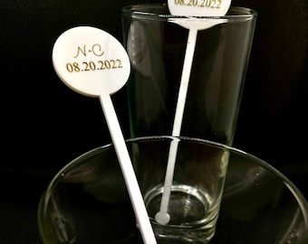 May include: Two white acrylic drink stirrers with round tops. The tops have gold lettering that reads "N+C 08.20.2022". The stirrers are in a clear glass and a clear glass bowl.