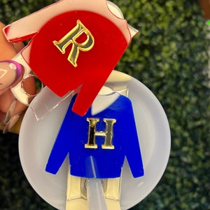 Puede incluir: Dos adornos de acrílico en forma de suéter con letras doradas. El suéter rojo tiene la letra "R" y el suéter azul tiene la letra "H". Los adornos están unidos a una tapa blanca con una pajita.