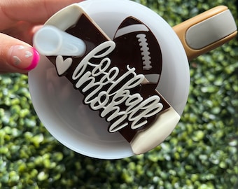 May include: A white ceramic mug with a brown and white football-themed design. The design features a football, a heart, and the words "football mom".
