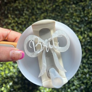 May include: White plastic cup lid with a white bow and the name "Brit" written on it. The lid has a hole for a straw.