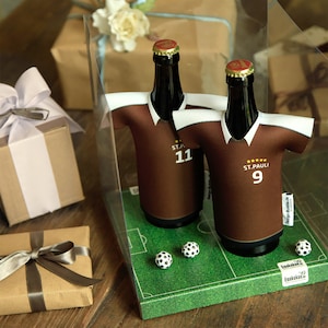 Könnte beinhalten: Zwei braune Bierflaschen mit braunen und weißen Trikot-Flaschenkühlern. Die Trikots tragen den Text "St. Pauli" und die Nummern "9" und "11". Die Flaschen werden auf einer grünen Filzoberfläche mit einem Fußballfeld-Design und weißen Fußbällen präsentiert.