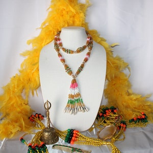 May include: A beaded necklace with a tassel and shell accents. The necklace is displayed on a white mannequin with yellow feather boas on either side. There are also several beaded necklaces and a small bell on the table.