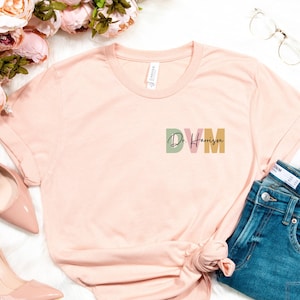 May include: A light pink t-shirt with a graphic design that says "DVM Dr. Harrison" in colorful letters. The shirt is laid out on a white surface with a pair of pink high heels, a pair of jeans, and a bouquet of pink flowers.