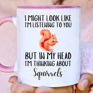 May include: White ceramic mug with a pink handle and interior. The mug features a watercolor illustration of a squirrel and the text: "I MIGHT LOOK LIKE I'M LISTENING TO YOU BUT IN MY HEAD I'M THINKING ABOUT Squirrels."