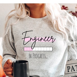 May include: A light gray sweatshirt with the word "Engineer" in black script and a pink progress bar graphic. The text "IN PROGRESS..." is below the graphic. A black mug is held in the hand.