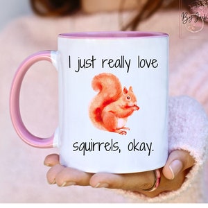 May include: White ceramic mug with a pink rim and handle. The mug has a watercolor illustration of a squirrel and the text "I just really love squirrels, okay."