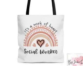 Puede incluir: Bolsa de tela blanca con asas negras. La bolsa presenta un diseño de arcoíris con corazones rosas y marrones y el texto "It's a work of heart Social Worker".